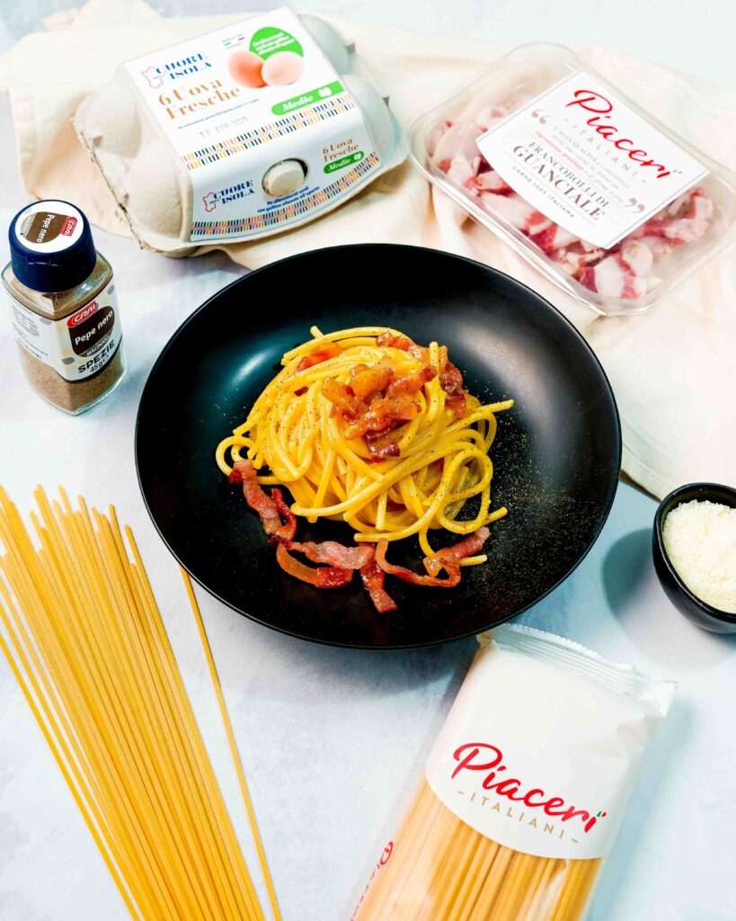 piatto nero con la pasta tipologia carbonara formaggio nella ciottola, confezione di pasta sul tavolo aperta e pepe e uova e scatolina di guanciale