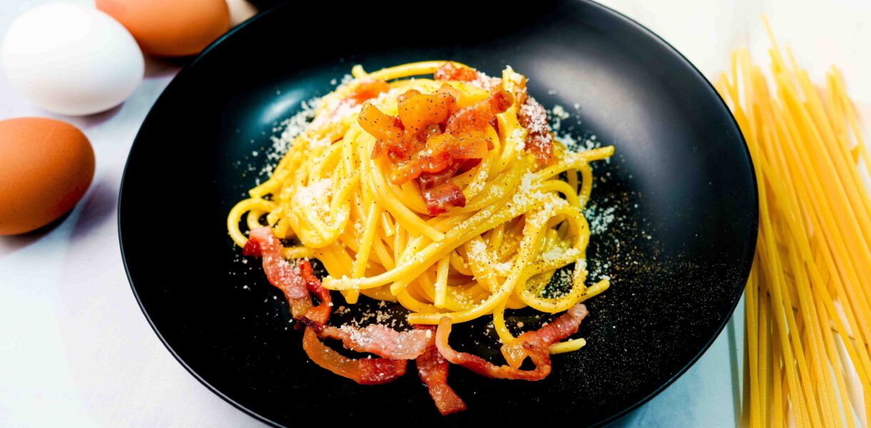 piatto di pasta alla carbonara nero, sul tavolo bianco con uova e spaghettoni e formaggio
