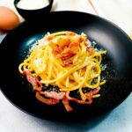 piatto di pasta alla carbonara nero, sul tavolo bianco con uova e spaghettoni e formaggio