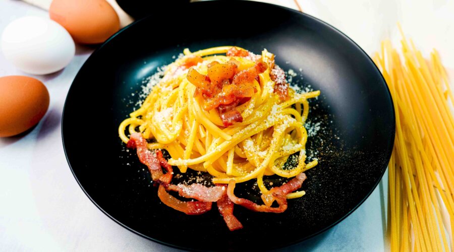 piatto di pasta alla carbonara nero, sul tavolo bianco con uova e spaghettoni e formaggio