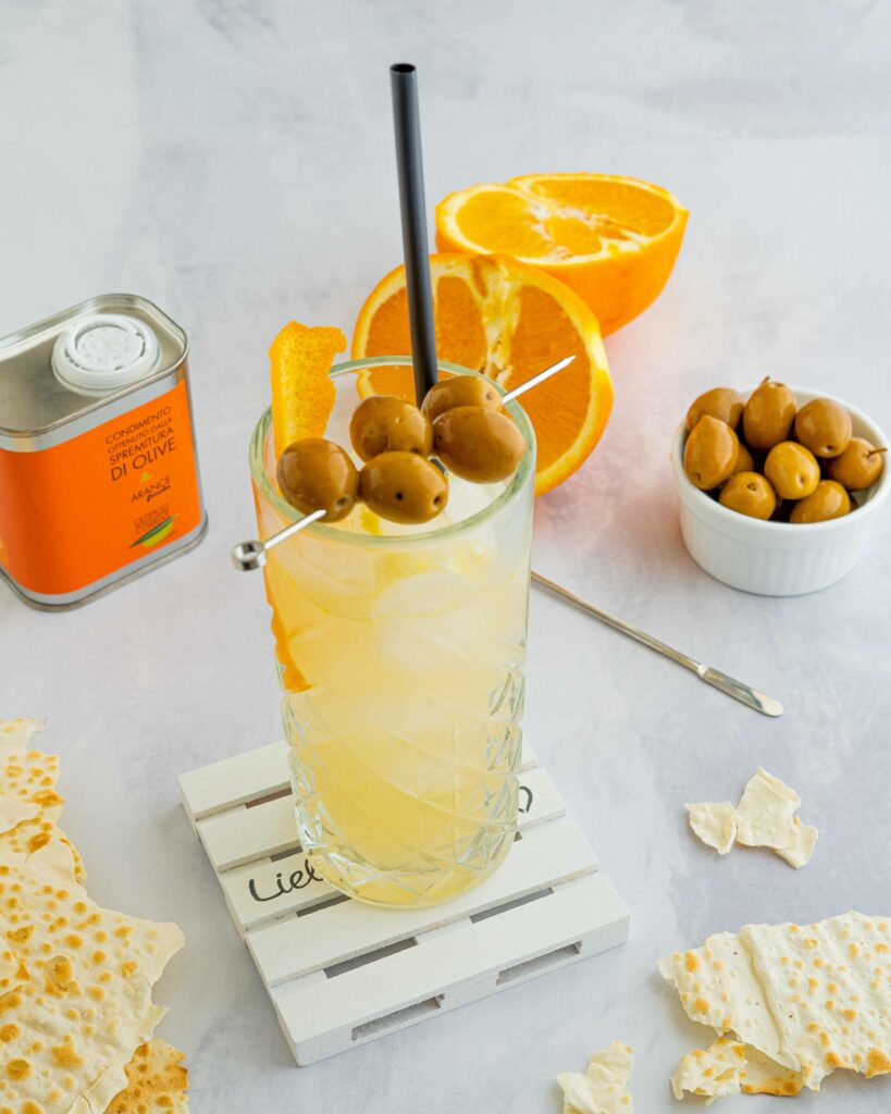 cocktail nel bicchiere trasparente con olive infilzate sopra, scorza di arancia, sul tavolo pane musica e arancia tagliata a metà. nel fondo latta di olio.