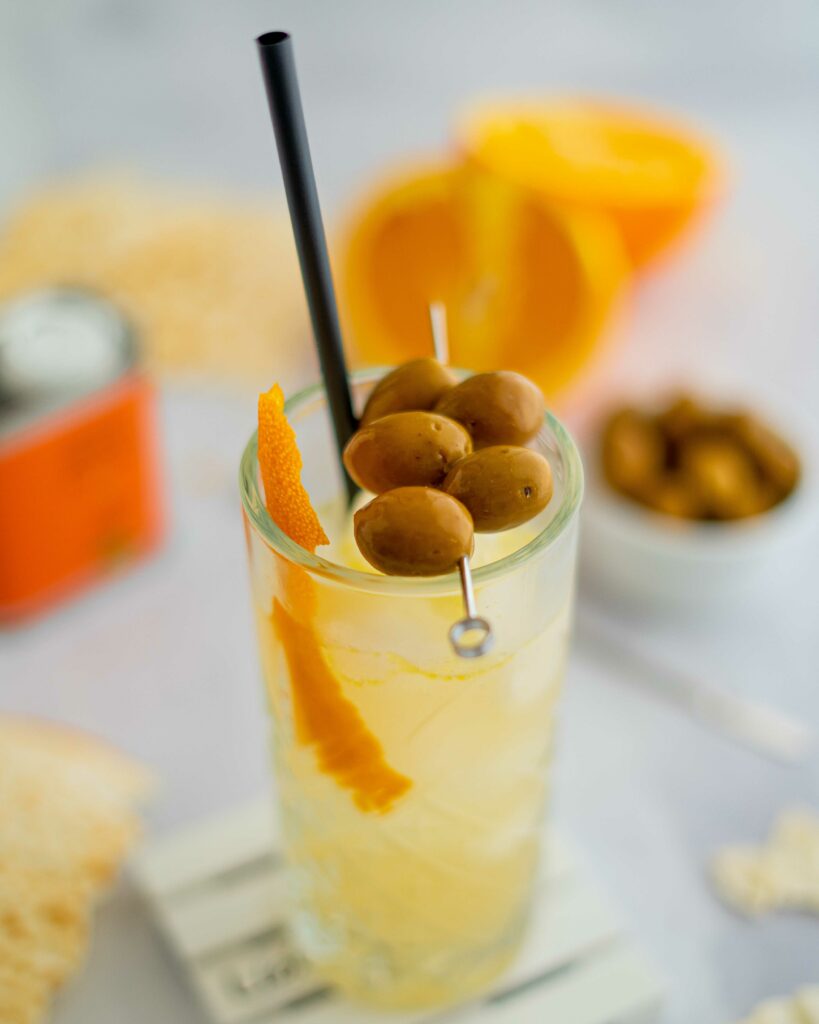 cocktail nel bicchiere trasparente con olive infilzate sopra, scorza di arancia, sul tavolo pane musica e arancia tagliata a metà con cannuccia.