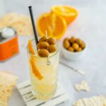 cocktail nel bicchiere trasparente con olive infilzate sopra, scorza di arancia, sul tavolo pane musica e arancia tagliata a metà.