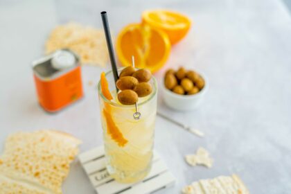cocktail nel bicchiere trasparente con olive infilzate sopra, scorza di arancia, sul tavolo pane musica e arancia tagliata a metà.