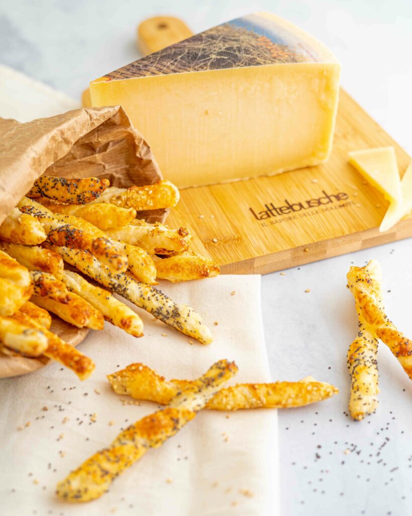formaggio sul tagliere intero a spicchio, nella busta di carta grissini sullo strofinaccio bianco, nel tavolo qualche grissino.