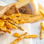 grissini dentro una busta di carta aperta con formaggio nel tagliere e sul tavolo qualche grissino
