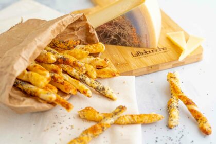 grissini dentro una busta di carta aperta con formaggio nel tagliere e sul tavolo qualche grissino