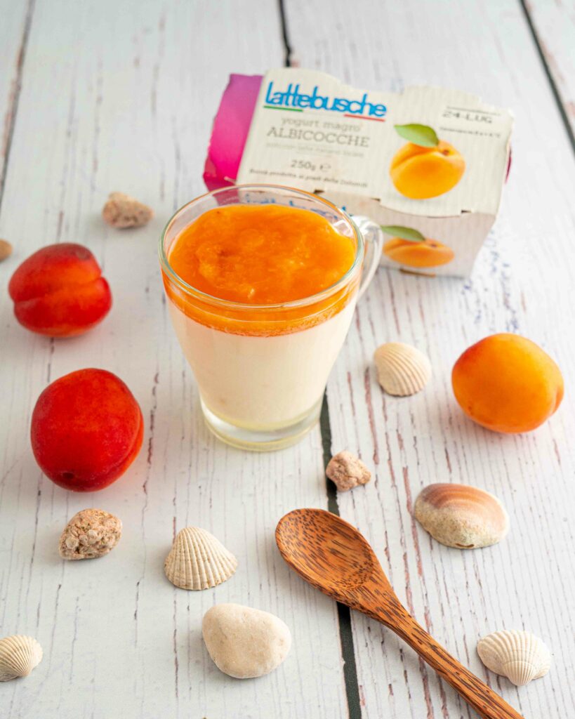 bicchiere con il budino cucchiaino sul tavolo con albicocche e conchiglie e scatola di yogurt