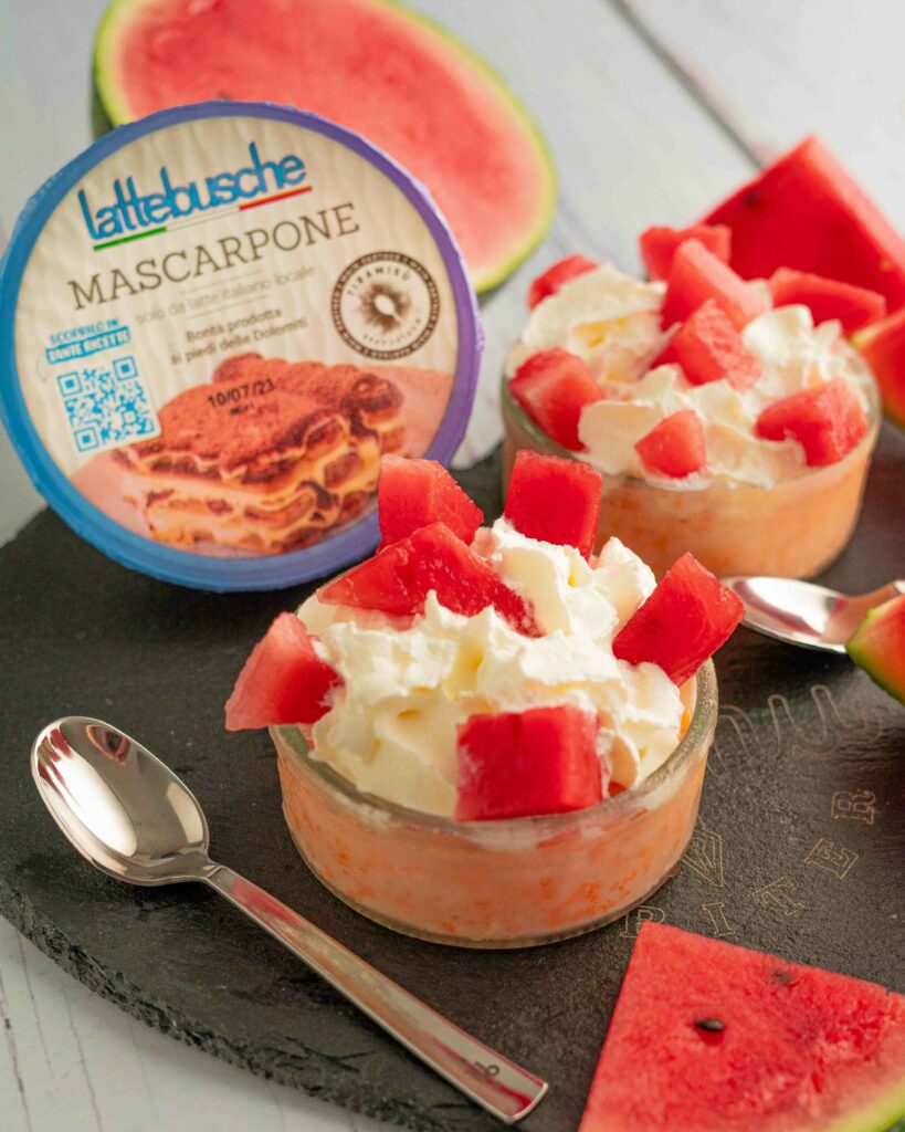 ciotola trasparente in primo piano con semifreddo e anguria a pezzi, confezione di mascarpone e cucchiaino sul tagliere