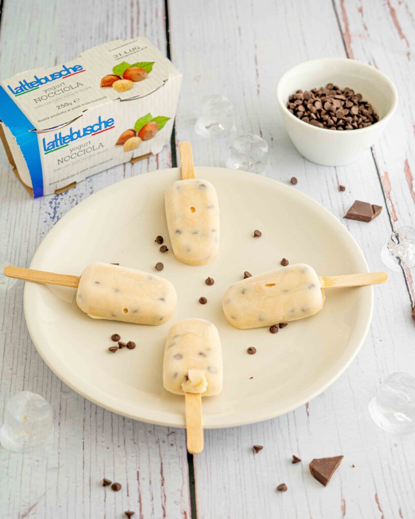 ghiaccioli sul piatto con gocce di cioccolato, sul tavolo ciotola con gocce di cioccolato, pezzi di cioccolato e cubetti di ghiaccio, in fondo confezione di yogurt.