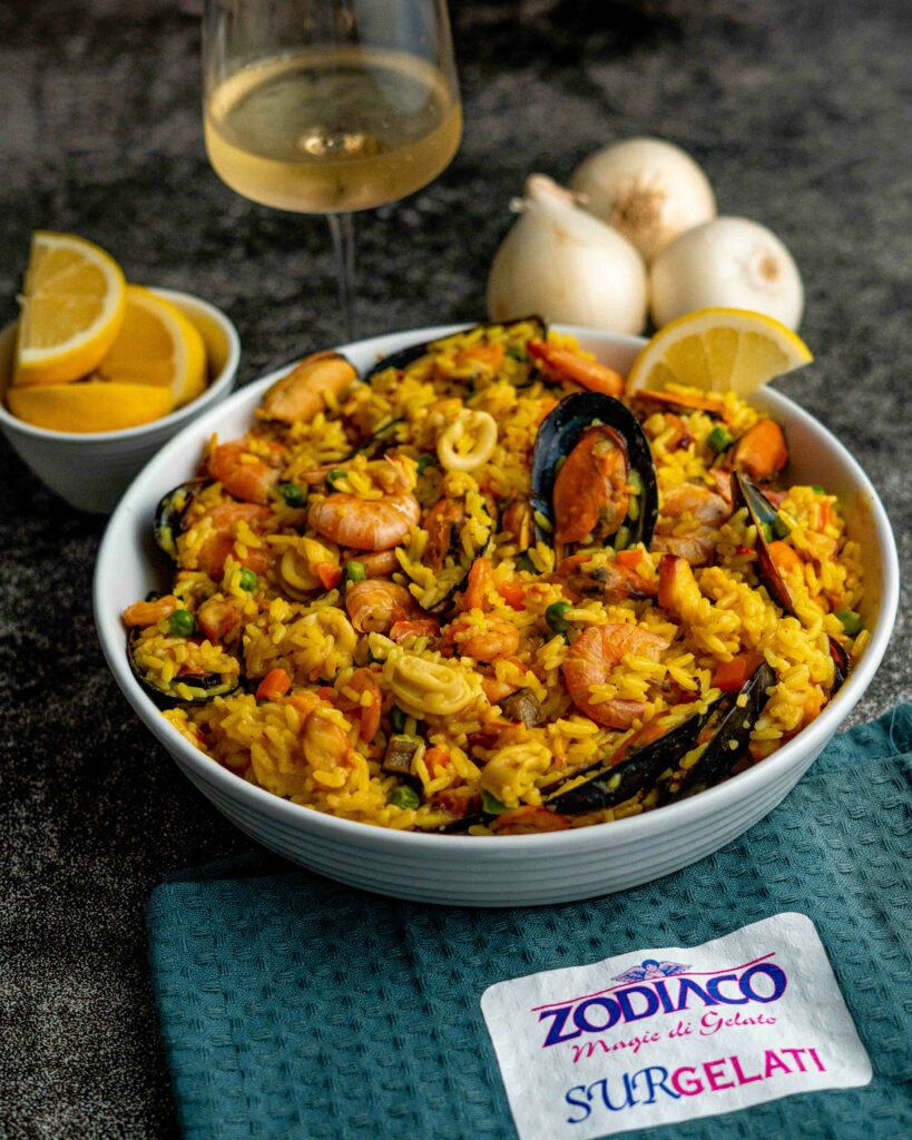 PAELLA DI PESCE SUL CONTENITROE CON LIMONE E CIPOLLE SUL TAVOLO  E BICCHIERE DI VINO BIANCO
