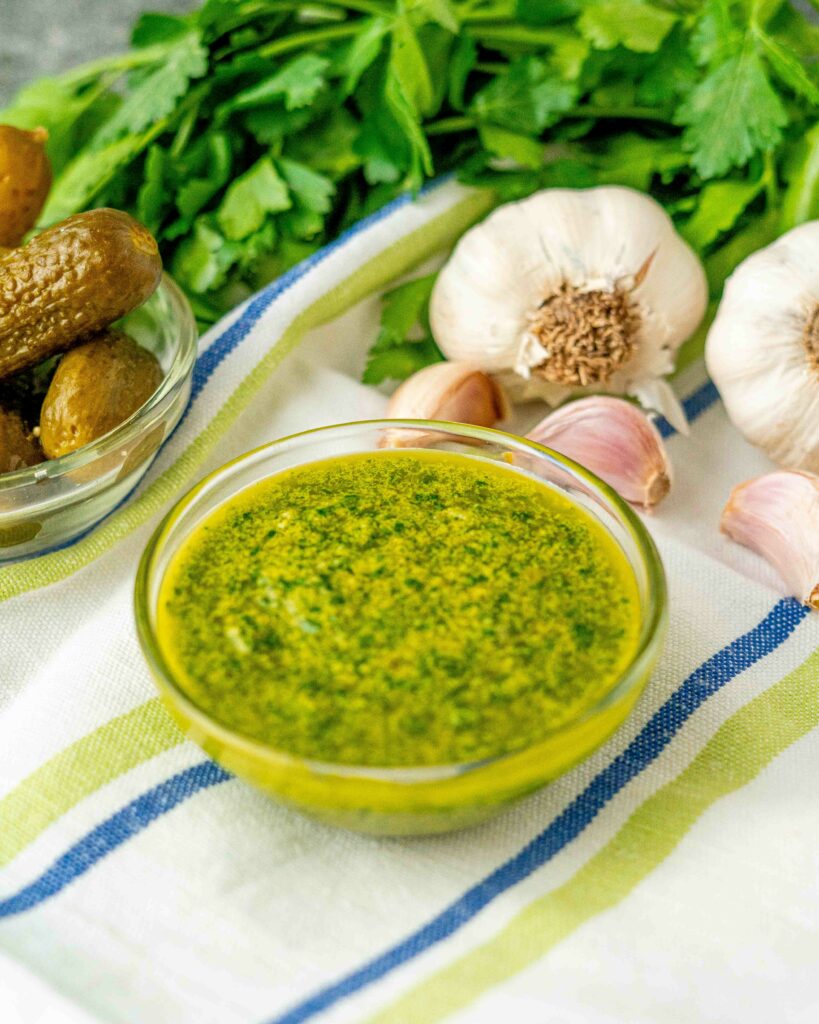 ciotola con salsa verde in primo piano con teste di aglio e spicchi di aglio e mazzo di prezzemolo sullo strofiaccio